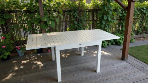 Table de repas extensible EOS 90 en aluminium beige, idéale pour les repas en extérieur, avec une extension de 90 à 180 cm, sur une terrasse en bois avec jardin en arrière-plan.