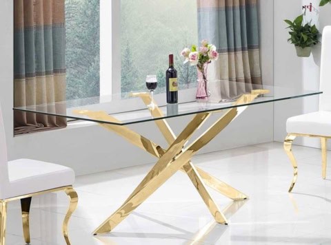 Alt text: Table à manger design avec plateau en verre et structure en métal doré, dans une pièce lumineuse avec fenêtres et végétation extérieure visible derrière.