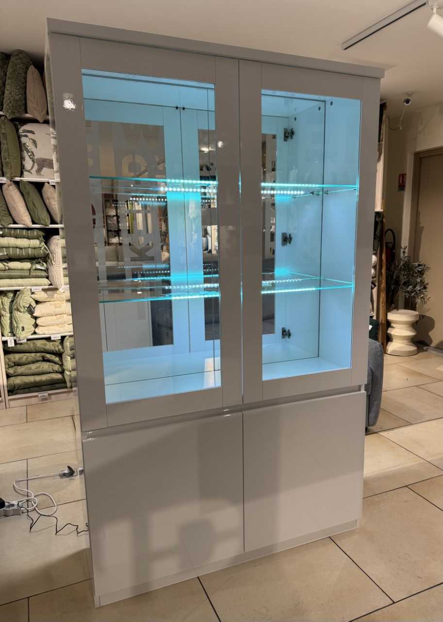 Vitrine moderne avec deux portes vitrées éclairées par LED, finition blanc laqué, offrant un espace de rangement inférieur pratique. Parfait pour exposer des objets dans un style élégant et contempora.